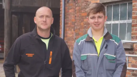 Ian Norbury A man and a boy standing in farming overalls. 