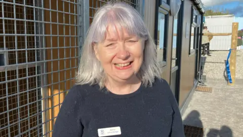A picture of Beverly smiling at the camera. She is wearing a black jumper and has long grey hair.