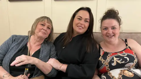 Three women smile while sitting on a settee indoors. One has a bob length mousey brown hair with a fringe, she is wearing a grey cardigan and has a tattoo on her arm, one is wearing black and has a gold bracelet and dark long hair. One has curly hair in an up-do with gold ear-rings - she is wearing a patterned dress with red and gold chains