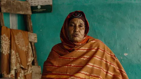 Netflix Saida Omar Mohamud in an orange and gold striped headscarf is talking and looking at the camera. She is sitting in front of a green wall.