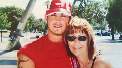 An archive picture of Michael Porter with his mother Jean Halon. He is wearing a red baseball cap and a red sleeveless tshirt. She has shoulder-length brown hair and is wearing sunglasses.