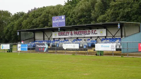 SHAUN WHITMORE/BBC Kirkely and Pakefield Football Club "Huggy" Stand, named after James Hutchinson. It has a row of seats five deep in blue and white colours and there are various advertising boards attached to a metal framed fence in front of the corrugated iron stand. You can see the green pitch in front of it.