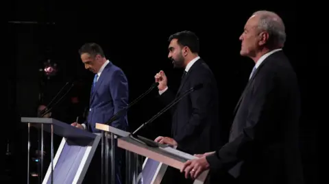 (l to right) Andrew Cuomo, Zohran Mamdani and Curtis Sliwa appear on a darkened stage in profile,  each behind a podium. They are wearing suits and Mamdani is speaking with one hand raised. 