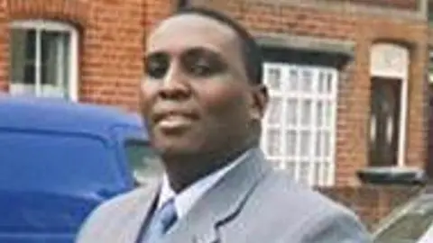 A grainy picture of Kevin Taylor stood looking at the camera in a grey suit and silver tie. He is standing in front of a blue van and houses.