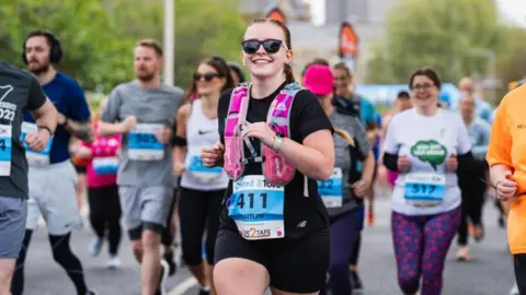 A runner wearing sunglasses, black t-shirt, black shorts and a pink hydration vest smiles into the camera as she takes part in the Grimsby 10K. A number of other runners are visible in the background.