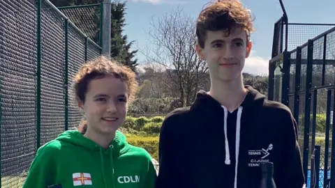 Two individuals standing on a tennis court holding trophies and water bottles. The person on the left is wearing a green hoodie with the text 'HISCOX' and 'CDLM' and a Guernsey flag printed on it. The person on the right is wearing a black hoodie with 'CI JUNIOR TENNIS' printed on it.