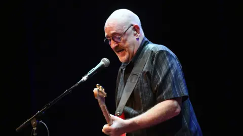  Scott Dudelson/Getty Images Dave Mason in a dark striped shirt with a red guitar strapped over his shoulder - he plays the guitar and sings into a microphone
