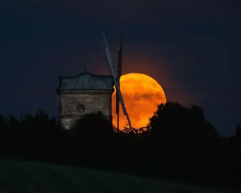BBC Weather Watchers / IanP A bright orange, red Moon is illuminated behind the sails of a windmill with trees outlined against the sky.