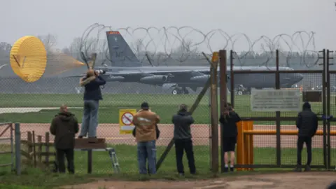 Reuters People watch from behind a barbed wire fence at the the U.S. Air Force Boeing B-52 Stratofortress bomber landing at RAF Fairford airbase, used by United States Air Force (USAF) personnel, amid the U.S.-Israeli conflict with Iran,