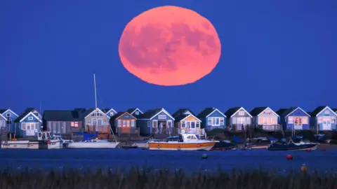 Andrew Wells Photography A row of large beach huts. Boats are in the water in the foreground. The moon is above, large, orange, a little elliptical. 