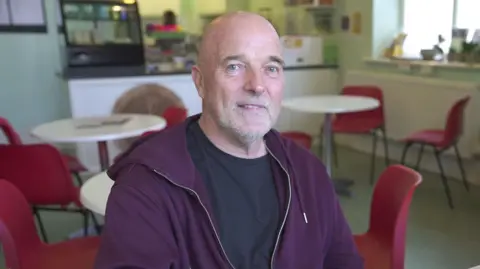Ian Mickiewicz in the old miner's hospital. It is a head and shoulders shot. He is sat in a cafe and the bar can be seen behind him, there are white tables and red chairs. Ian is a bald man with a stubbly beard and moustache. He has a black t-shirt on and over that a purple hoody that is unzipped. He is looking at the camera.