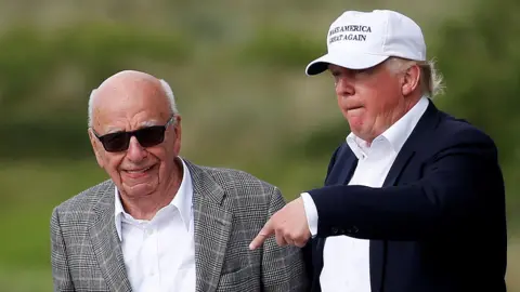 Rupert Murdoch, who wears dark sunglasses and a check suit, smiles as he walks with Donald Trump, who wears a suit and a MAGA cap