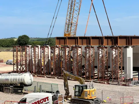 A large bridge structure made of brown steel beams on a building site. It has not yet been lifted into place but is surrounded by construction vehicles including vans, a tanker and a yellow digger.
