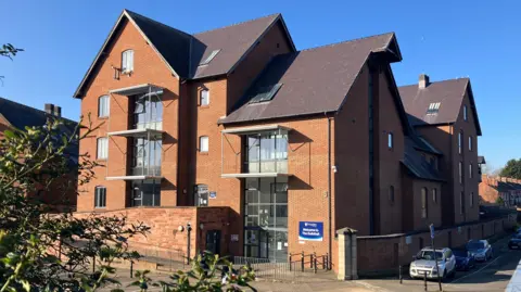 A brown brick building with five storeys and large glass windows. There is a blue and white sign next to a glass entrance door that reads "Welcome to The Guildhall"