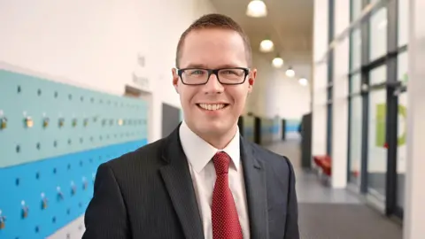 Ian Loynd Ian Loynd, headteacher at St Teilo's Church in Wales High School, Cardiff, photographed in the halls of the school. He wears a black suit, pale pink shirt and red and white polka dot tie. He wears black rectangle glasses and smiles at the camera.