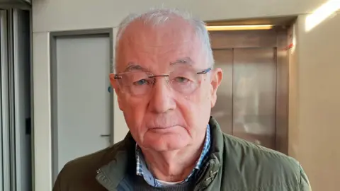 Mickey McKinney has short grey hair. He is wearing glasses and has a green coat on with a blue shirt and jumper. He is standing in a reception area with a lift in the background. 