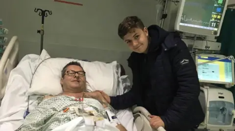 Jack Rondel pictured with his father Richard in hospital. Richard is lying in bed, while Jack is stood bedside in a black coat. They are both smiling, Richard is wearing black glasses. hospital equipment is visible in the background.