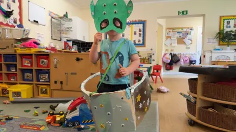 A little boy wearing a home-made costume of an alien with a green mask and a silver spaceship around his waist. He is in a nursery classroom, surrounded by toys.