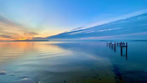 Most of the shot shows a wide expanse of water with wooden groynes on the right-hand side. Both the water and the sky are a very vivid blue colour that darkens from left to right. The first rays of the sun also create a golden glow on the horizon on the left-hand side of the picture.