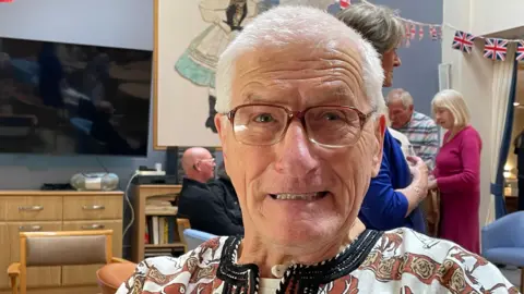 Charles Jagla. He has short grey hair and glasses. He is wearing an African style shirt with safari style animals and patterns. 