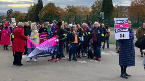 A woman in a long blue coat holds a placard reading 'enough is enough' while a group of women gather behind holding banners that say reclaim the night. They are wearing yellow arm bands.