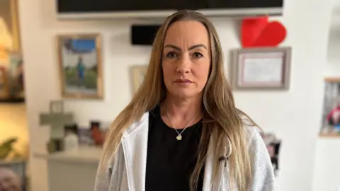 Linzi Stewart is in her lounge, standing in front of a fire place, with a photo of her brother Tim on a table nearby