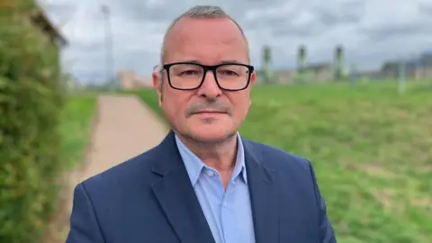 Sam Read/BBC Lee Barron, wearing a blue shirt, blue suit jacket, standing outside, wearing black-rimmed glasses, looking straight at the camera. Grass and buildings are behind him, which is blurred. 