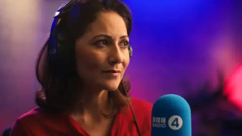 BBC Mishal Husain wearing headphones and in front of a microphone, looking pensively into the distance against a blurred purple and blue background