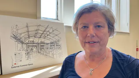 Sally pictured smiling slightly and looking into the camera. She has short grey hair and is wearing a navy blue top and a silver necklace. To her right is a mock-up drawing of the market after a refurb - it's drawn in pencil, like a sketch, and shows a food court.