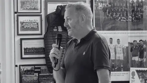 Birmingham & Solihull RFC A black and white photo of Matt Gallagher talking into a microphone, stood in an office with rugby memorabilia on the walls