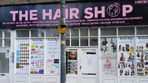 The front of a large shop with panelled windows. Numerous posters of hair and hair products can be seen advertised on the front.