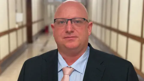 BBC Man (bald, glasses) in dark jacket, blue shirt, pink tie in long corridor of Derbyshire's county hall (high ceilings, dark and cream panelling on walls)