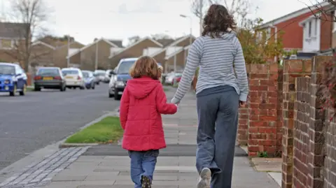 A young girl wearing a red coat is walking hand in hand with her mother on a suburban street