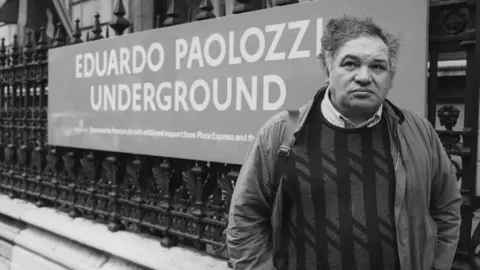 Hulton Archive via Getty Images A black and white photo of a middle-aged man with dark wave hair standing outside a building with decorative black railings and a large sign reading "Eduardo Paolozzi Underground". He is wearing a dark jacket over a jumper with vertical stripes and a light checked shirt.
