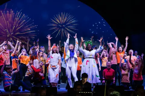 Elly Welford The finale of Sing, Dance, Leap with the three lead characters in white costumes surrounded by children in orange and fireworks in the background
