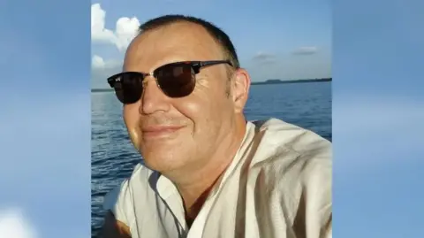 A man with short hair and sunglasses smiles while the setting sun shines in his face. Behind him is an expanse of open water and a strip of land in the far distance. He is wearing a cream shirt.