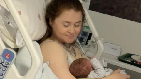 Stephen Whyte Lauren Evans in a hospital bed holding Ambrose. She is looking down at him. 