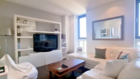 A photo of the inside of Amanda's flat which shows a widescreen TV inside a cabinet. A white l-shaped sofa and a coffee table sit in front of the TV with a window in the background. 