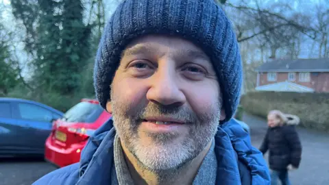 A man with a beard is smiling while wearing a blue knitted hat and a blue and grey coat while standing in front of a red car.
