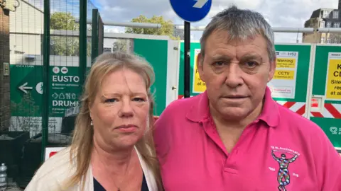 Karen and Craig Douglas standing in front of hoardings surrounding building site