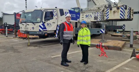 Matt Turmaine Matt Turmaine, wearing a white hard hat and orange and grey hi-vis, with black trousers and shoes, and Allison Phillips, wearing a blue hard hat and orange hi-vis with black trousers and shoes. They are standing in front of a white and blue lorry with a crane attached. A portable cabin is visible to the left of the picture.