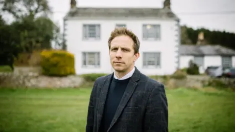 Hunter Hall Paul Borrows standing in a grassed area at the front of Hunter Hall school. He has dark hair, spiked at the front, and is wearing a dark blazer and navy jumper.