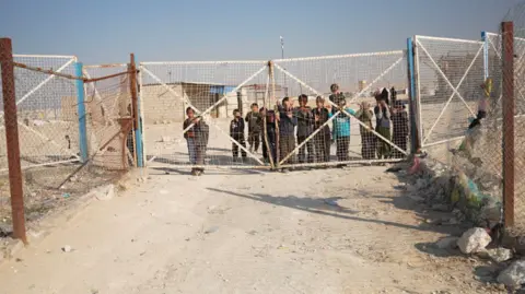 Children seen through a wire mesh fence at the end of a dirt track in the al-Hol camp in Syria