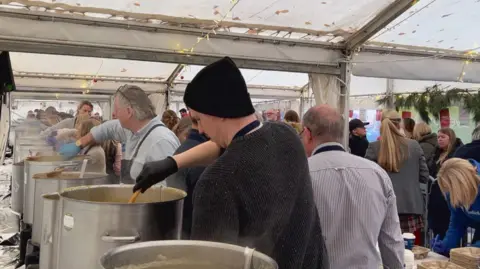 A busy food serving area inside a large tent, where several people are stirring and serving food from large metal pots placed on tables. Steam rises from the pots, and the setup includes utensils and containers for serving. In the background, more people are gathered, and the tent is decorated with string lights and greenery along the sides.