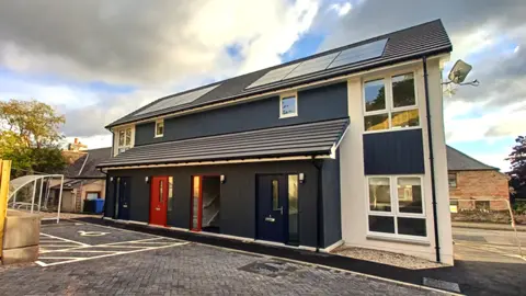 A modern looking building with four doors on the ground level, windows and solar panels on the roof. There's a car parkin area in front. A sunny and cloudy day.