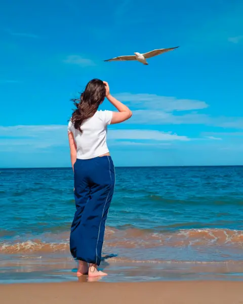 Nikki Smith A girl with brown hair, wearing a white T-shirt and blue trousers, holds her right hand to her head, with her back to the camera.
She is standing at the shore on a beach and a seagull is flying directly over her head, 