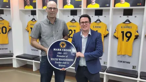 Two men stand in a football club's dressing room while holding a round blue plaque. The plaque says "Mark Crook, 1903-1977, founder of Wath Wanderers, feeder club to Wolves" underneath the yellow and black Wolves logo. Behind them are yellow Wolves players' shirts hung up on the dressing room wall.