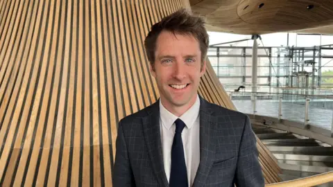 The image shows Sam Rowlands standing in front of the Senedd's funnel. He is looking at the camera and smiling. He is wearing a grey check suit, a white shirt and a navy tie.