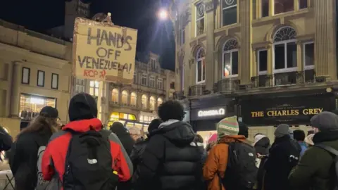 Emma Wass/BBC A protest by Newcastle Stop The War. A group of people in various coloured winter coats, hats and scarves are gathered around Grey's Monument in Newcastle City Centre. The sky above them is dark, and the streetlights/shop lights are lighting up the street. A home-made cardboard sign is being held up on a stick by one individual says 'hands off Venezuela!'.