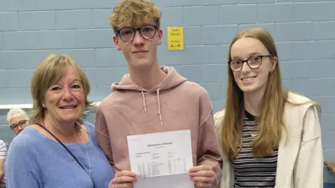 Supplied Beckett stands between his mum and sister holding his results, in a school hall. 